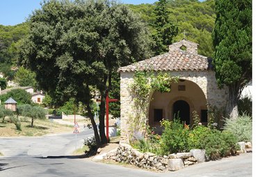 Entrée de la chapelle avec une ouverture voutée surplantée d'une petite croix en fer et entourée d'arbustes fleuris et d'arbres
